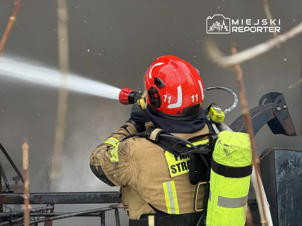 Strażak w hełmie z numerem 11 kieruje strumień wody z węża gaśniczego na płonący obiekt w zamglonym otoczeniu.