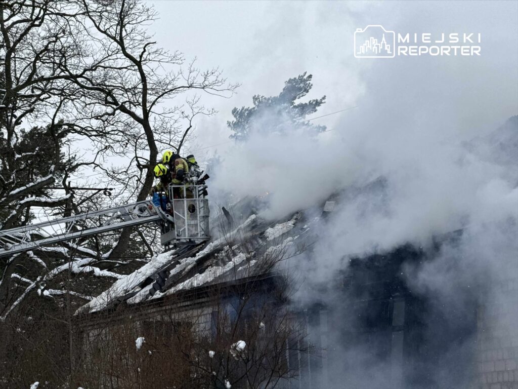 Strażacy w hełmach gaszą pożar na dachu budynku, otoczeni gęstym dymem i śniegiem.