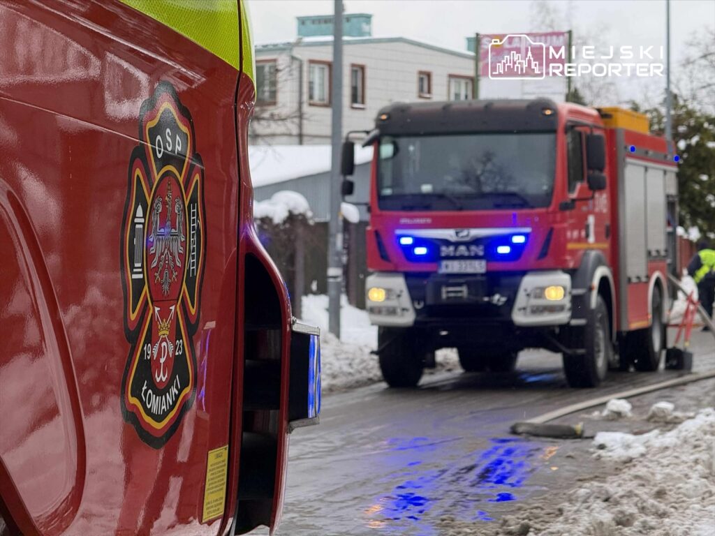 W tle widać wóz strażacki marki MAN z włączonymi sygnałami świetlnymi, a z przodu widoczny jest emblemat OSP Łomianki.