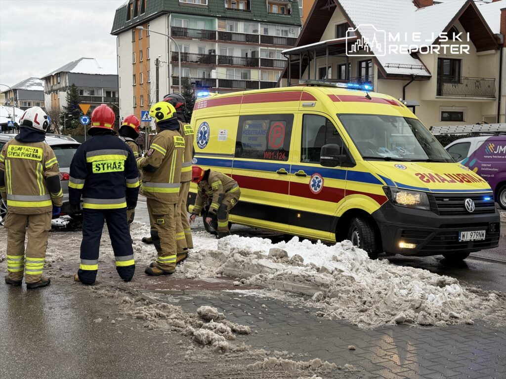 Grupa strażaków w mundurach, w tym jeden z hełmem, pomaga przy usuwaniu pojazdu z zaspy śnieżnej obok karetki.