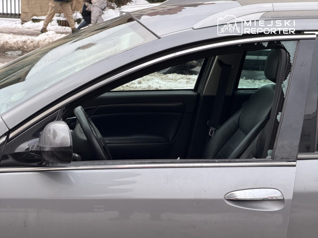 Uszkodzone auto z wybitym oknem, zaparkowane na chodniku w zaśnieżonej okolicy, obok przechodzący ludzie.