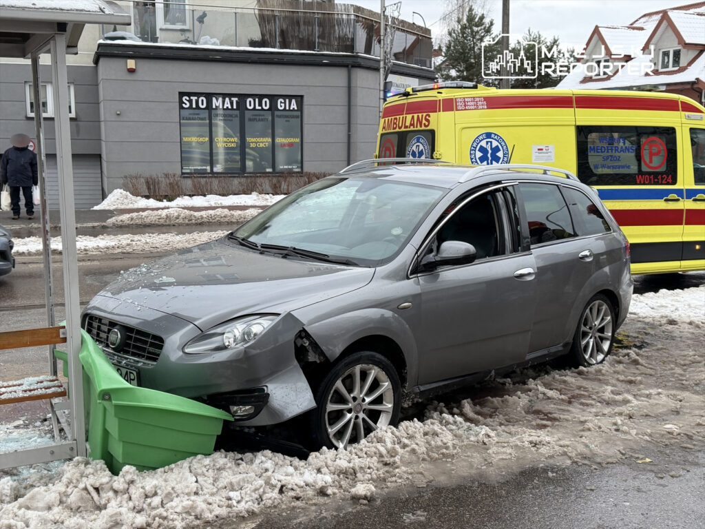 Samochód osobowy stoi na poboczu, z przodu uderzony w zielony pojemnik, obok leży szkło. W tle widać ambulans.