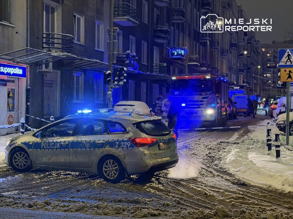 Policyjny radiowóz z włączonymi sygnałami świetlnymi stoi na zaśnieżonej ulicy, obok wozu strażackiego.
