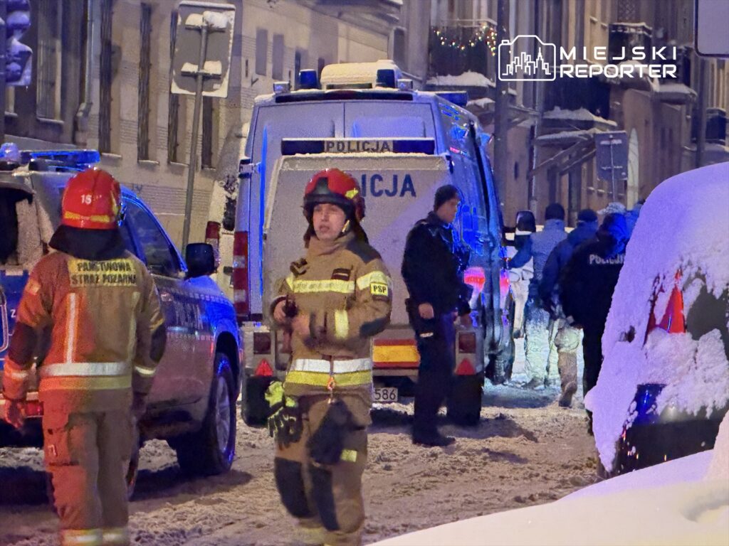 Grupa strażaków i policjantów prowadzi działania na zaśnieżonej ulicy, otoczonej budynkami.
