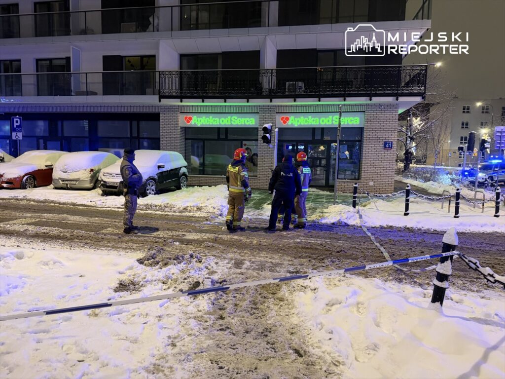 Grupa ratowników i policjantów rozmawia na zaśnieżonej ulicy przed apteką, otoczoną parkującymi samochodami.