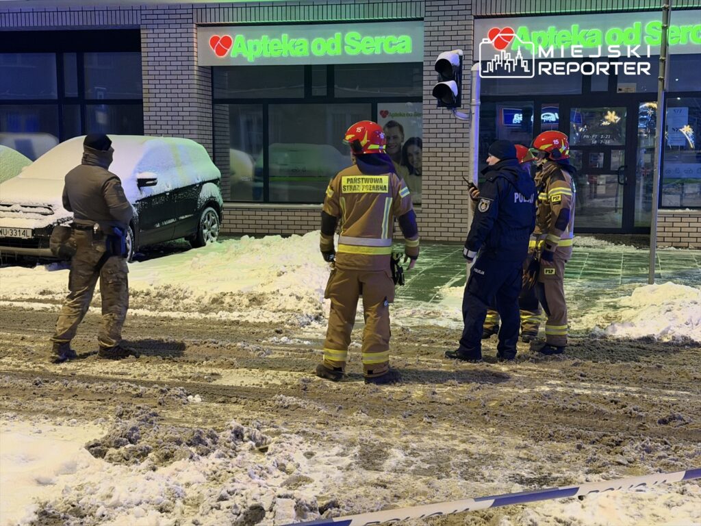 Czterej strażacy w mundurach rozmawiają z policjantem na zaśnieżonej ulicy przed apteką.