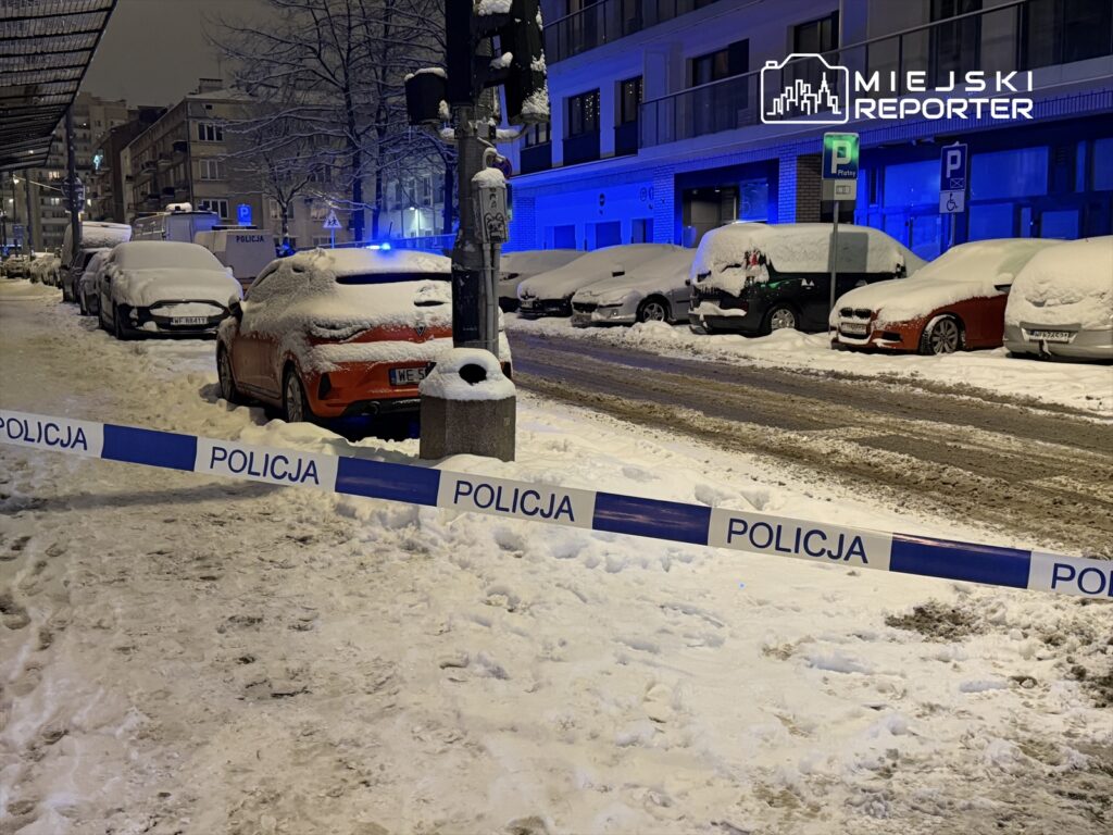Zasypana śniegiem ulica z zaparkowanymi samochodami, w tle niebieskie światła policyjnych radiowozów i taśma policyjna.