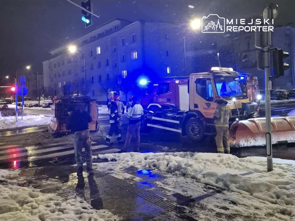 Grupa strażaków i pracowników miejskich zajmuje się przewróconą pługopiaskarką na ośnieżonej ulicy w nocy.