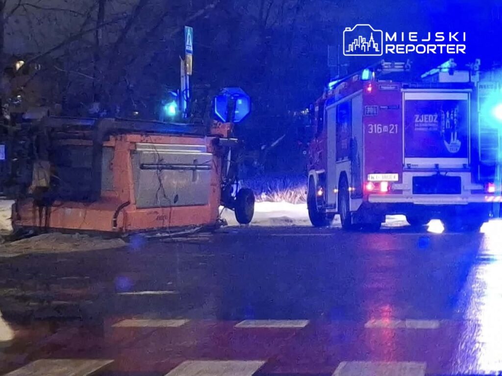 Przewrócony pojazd na drodze, obok straż pożarna z włączonymi sygnałami świetlnymi. Nocna sceneria z oświetleniem ulicznym.