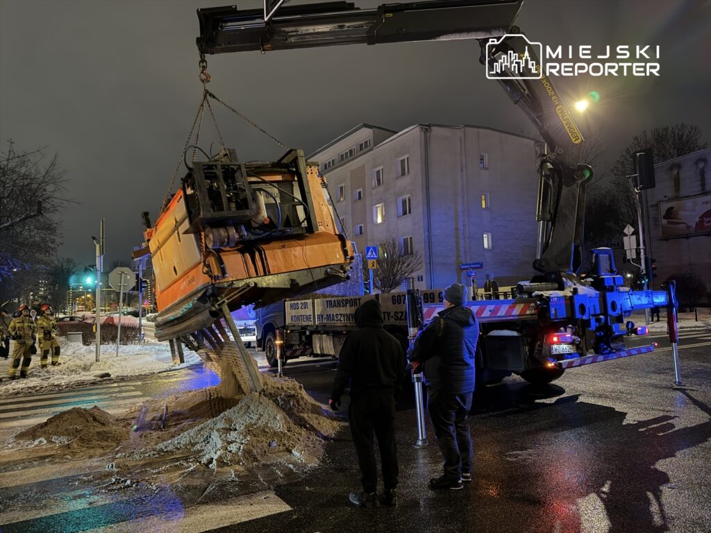 W nocy, w miejskim otoczeniu, podnośnik unosi uszkodzony pojazd, a obok stoją strażacy i obserwatorzy.