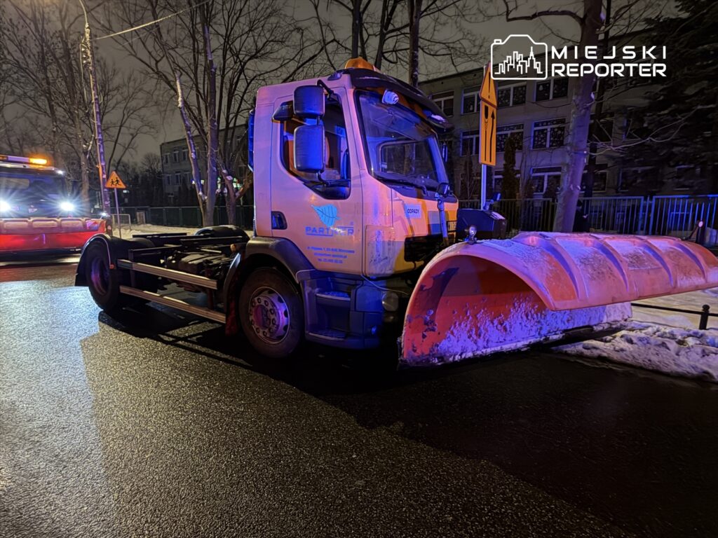 Pojazd z pługiem śnieżnym zatrzymany na oświetlonej ulicy, obok widoczny drugi pojazd ratunkowy.
