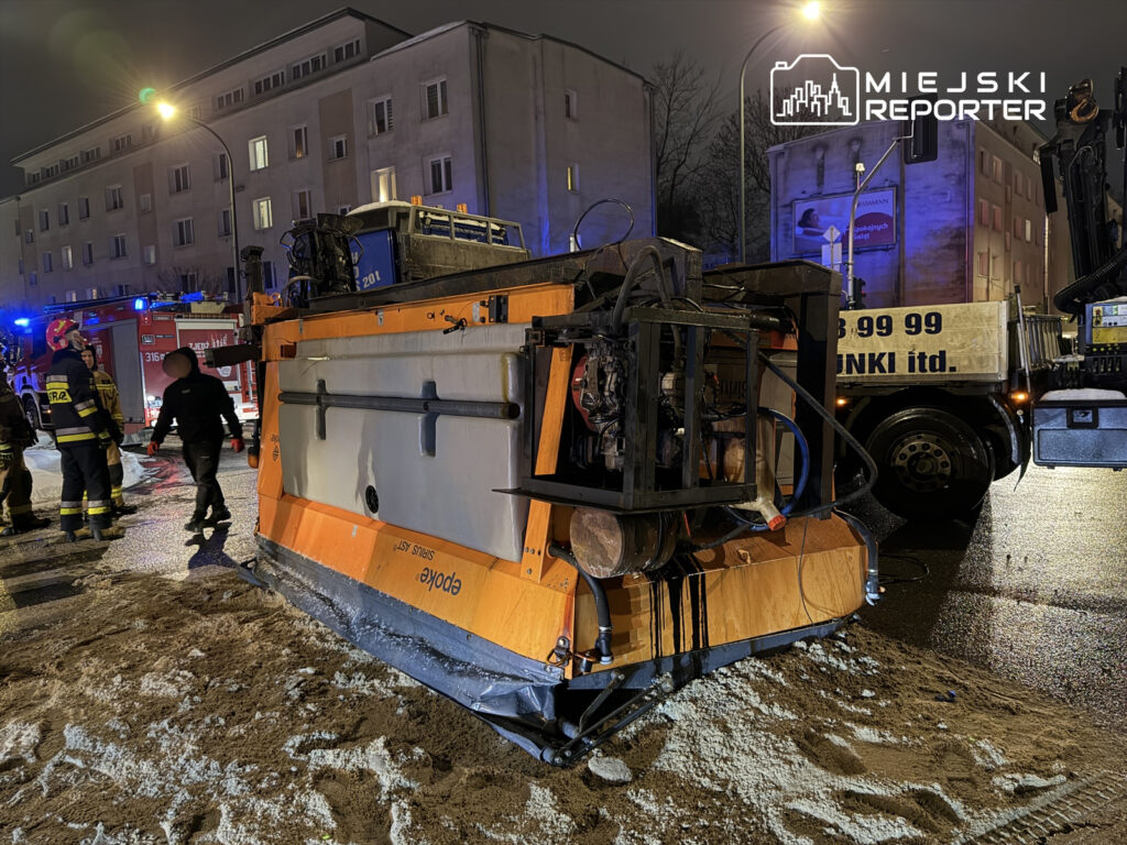 W nocy, strażacy pracują przy przewróconej maszynie budowlanej na ulicy, obok stoi samochód strażacki.