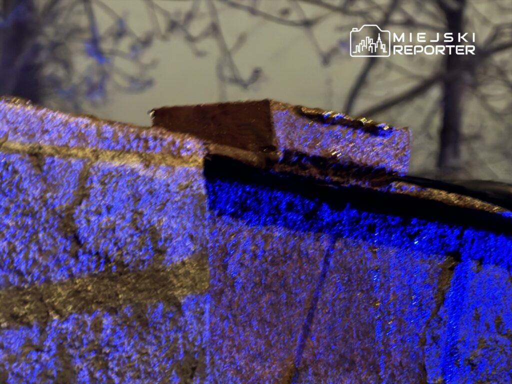 Kawałek metalowego elementu pokryty niebieskim światłem, z tłem przedstawiającym zamglony krajobraz drzew.