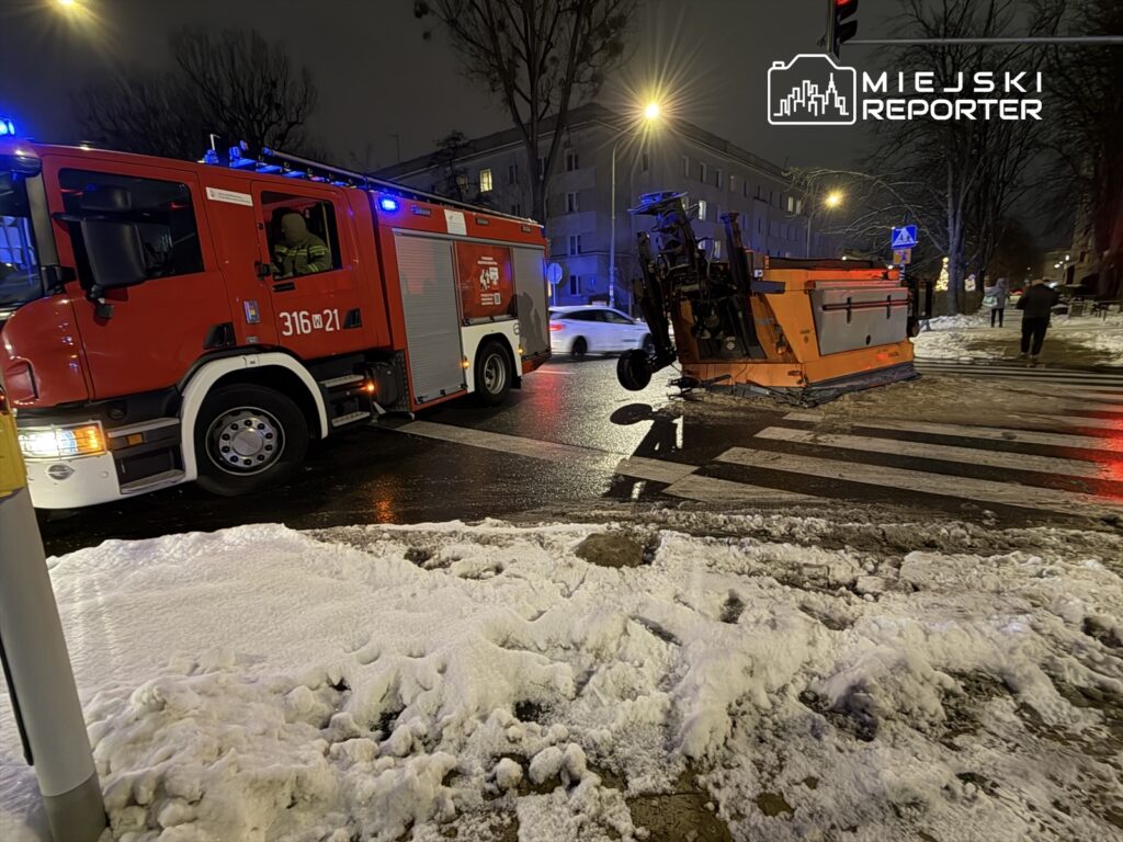Straż pożarna zatrzymana przy przewróconej maszynie do odśnieżania na ośnieżonej ulicy w nocy.