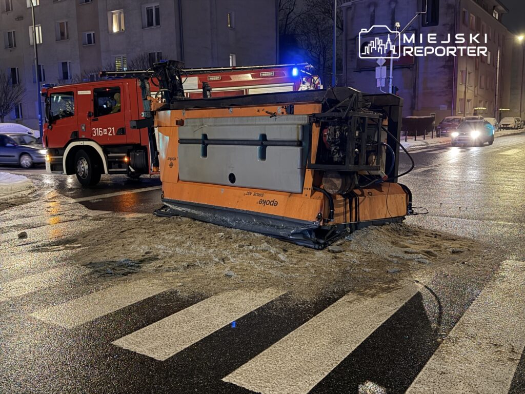 Wóz strażacki stoi obok przewróconej maszyny do odśnieżania na oświetlonej ulicy.