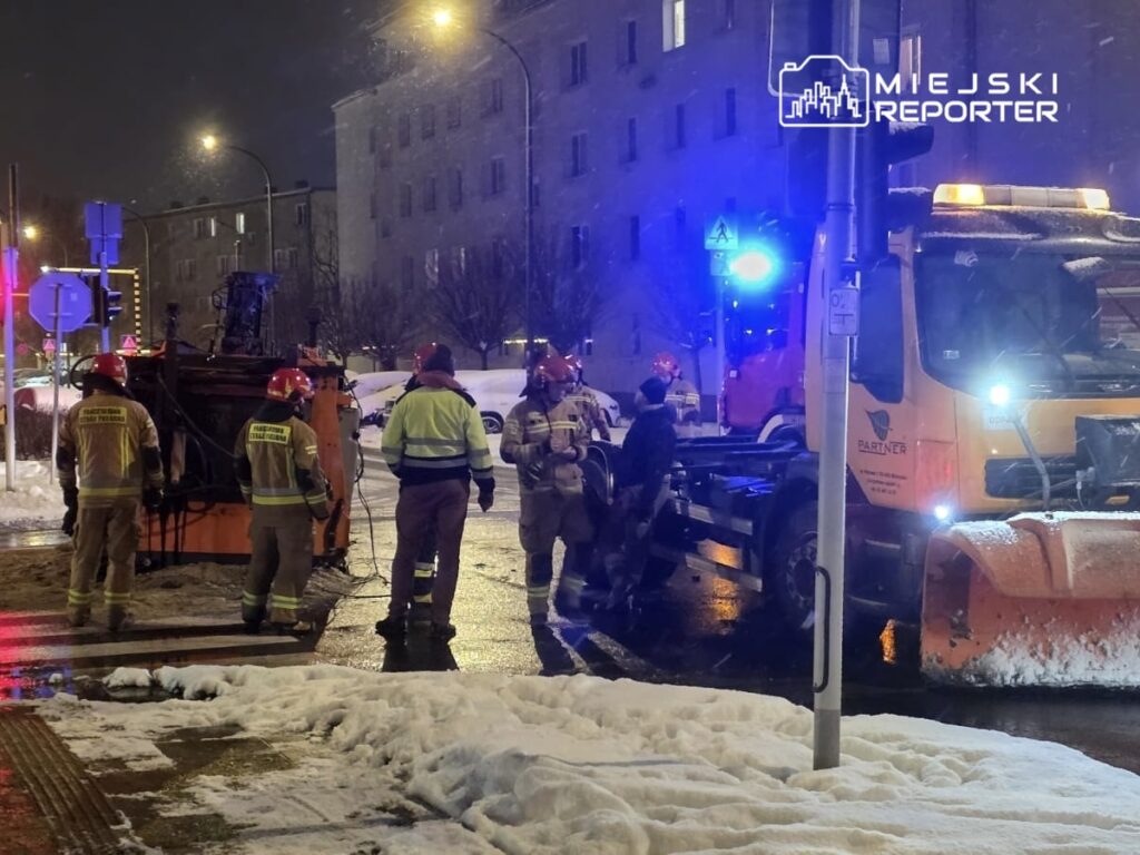 Grupa strażaków w odblaskowych mundurach pracuje przy przewróconej maszynie do odśnieżania na ośnieżonej ulicy nocą.