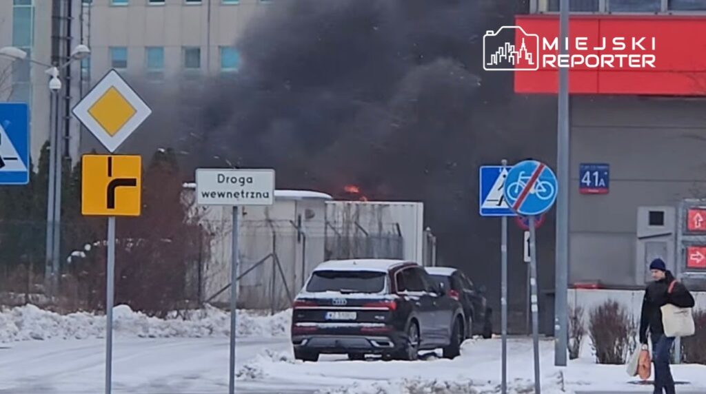 Gęsty czarny dym unoszący się z kontenera, obok zaparkowane samochody i znaki drogowe, w zimowej scenerii.