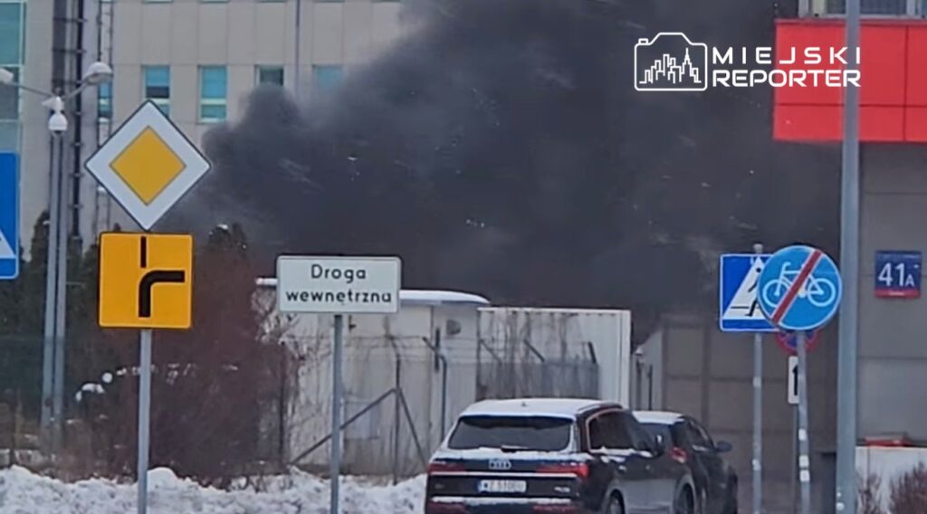 Czarny dym wydobywający się z budynku w miejskim otoczeniu, w tle znaki drogowe i samochody zaparkowane na śniegu.