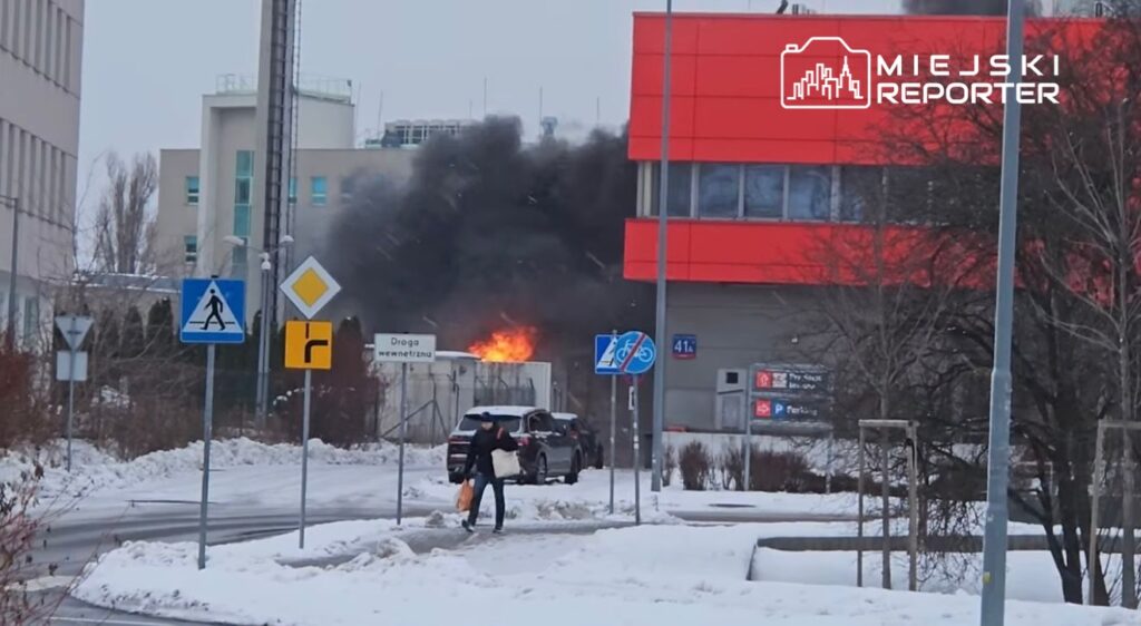 Mężczyzna z torbą przechodzi obok płonącego kontenera, z którego wydobywa się czarny dym w zimowej scenerii.