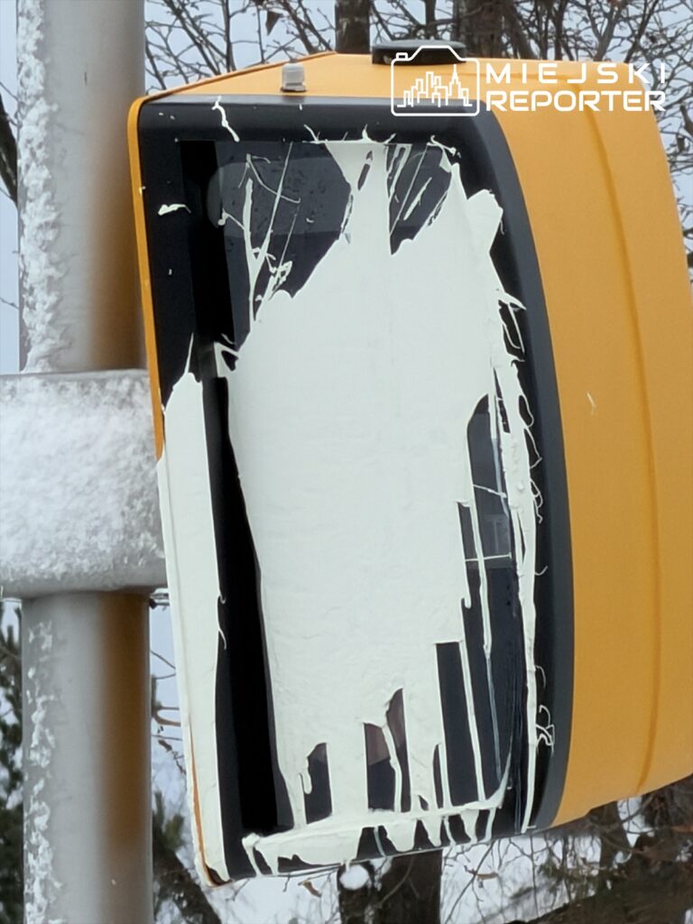 Zniszczony sygnalizator świetlny z białą farbą na obudowie, umieszczony na stalowym słupie w zimowym otoczeniu.