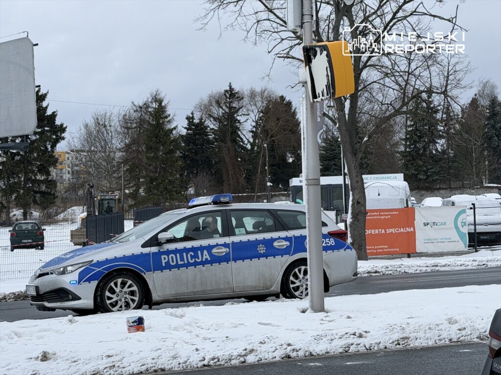 Policyjny radiowóz zaparkowany na ulicy w zimowej scenerii, obok zasp śnieżnych i billboardu.