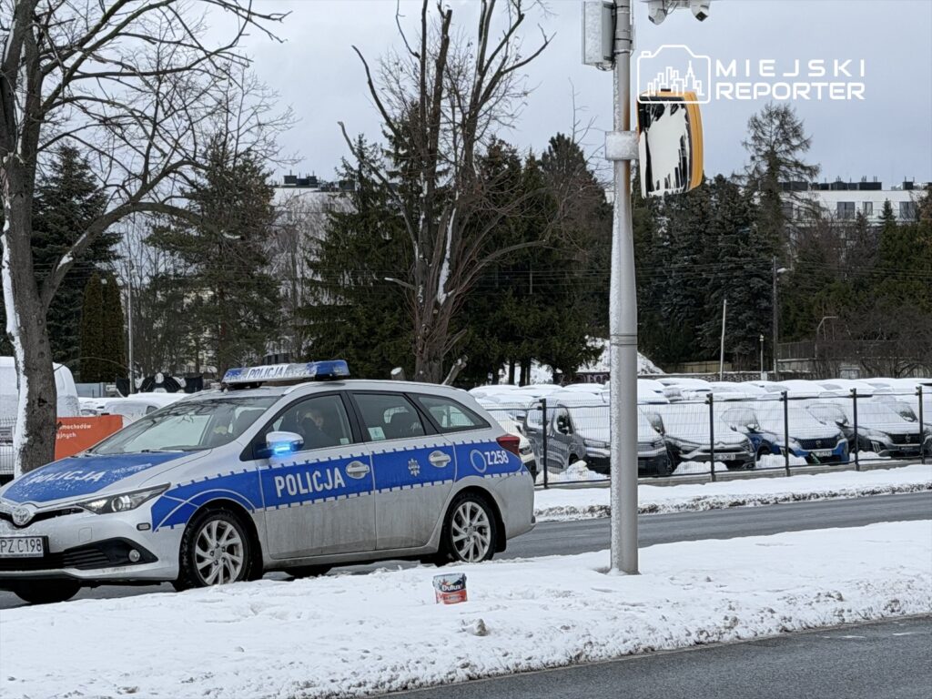 Policyjny radiowóz z włączonymi sygnałami świetlnymi stoi na zaśnieżonej ulicy obok parkingu z przykrytymi śniegiem samochodami.