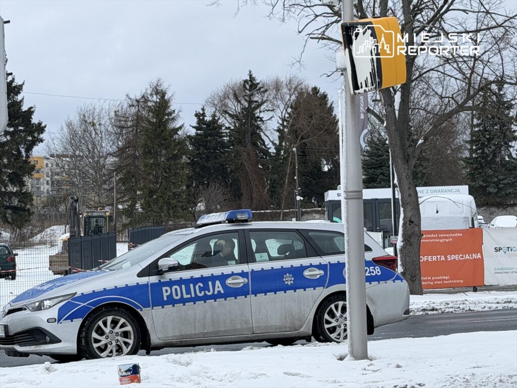 Policyjny radiowóz z niebieskimi światłami parkuje na zaśnieżonej ulicy, w tle widoczne drzewa i budynki.
