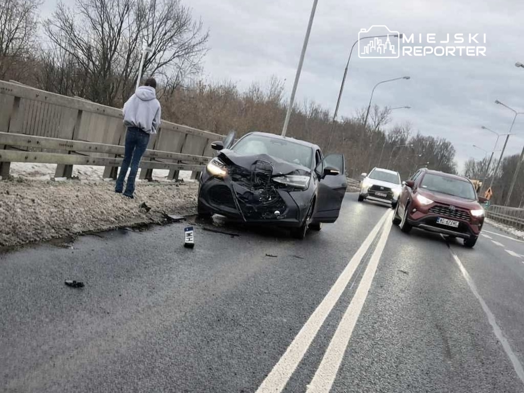 Młody mężczyzna w szarej bluzie stoi obok uszkodzonego samochodu na drodze, obok przejeżdżające inne pojazdy.