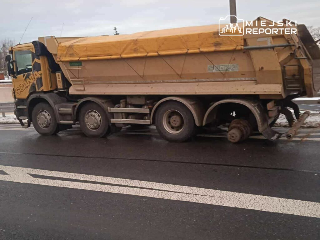 Żółty samochód ciężarowy marki Scania stoi na drodze, z uszkodzoną osią i widocznym brakiem jednego koła.