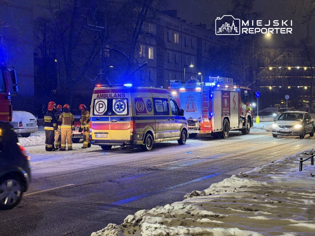 Grupa strażaków w mundurach omawia sytuację obok ambulansu i wozu strażackiego na ośnieżonej ulicy w nocy.
