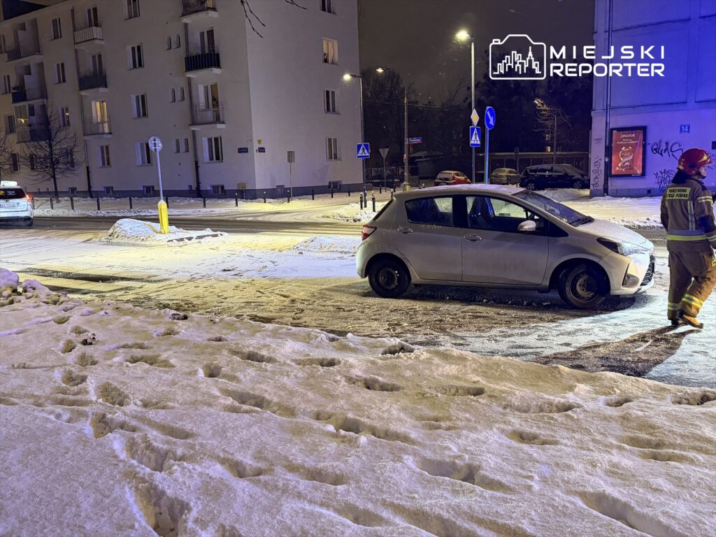 Samochód osobowy zaparkowany na zaśnieżonej ulicy, obok stojący strażak w żółtej kurtce.