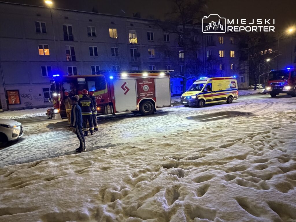 Strażacy w mundurach rozmawiają obok wozu strażackiego na zaśnieżonej ulicy w nocy. W tle widoczna karetka.