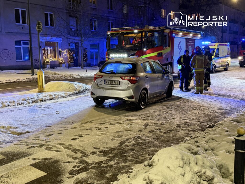 Na oświetlonej ulicy widać srebrny samochód zaparkowany obok grupy ratowników w mundurach, rozmawiających z osobami w pobliżu.