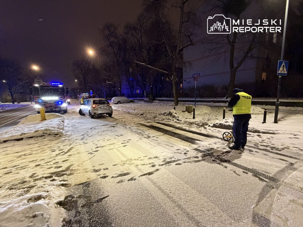 Policjant w odblaskowej kamizelce mierzy ślady na zaśnieżonej drodze obok uszkodzonego samochodu i wozu strażackiego.