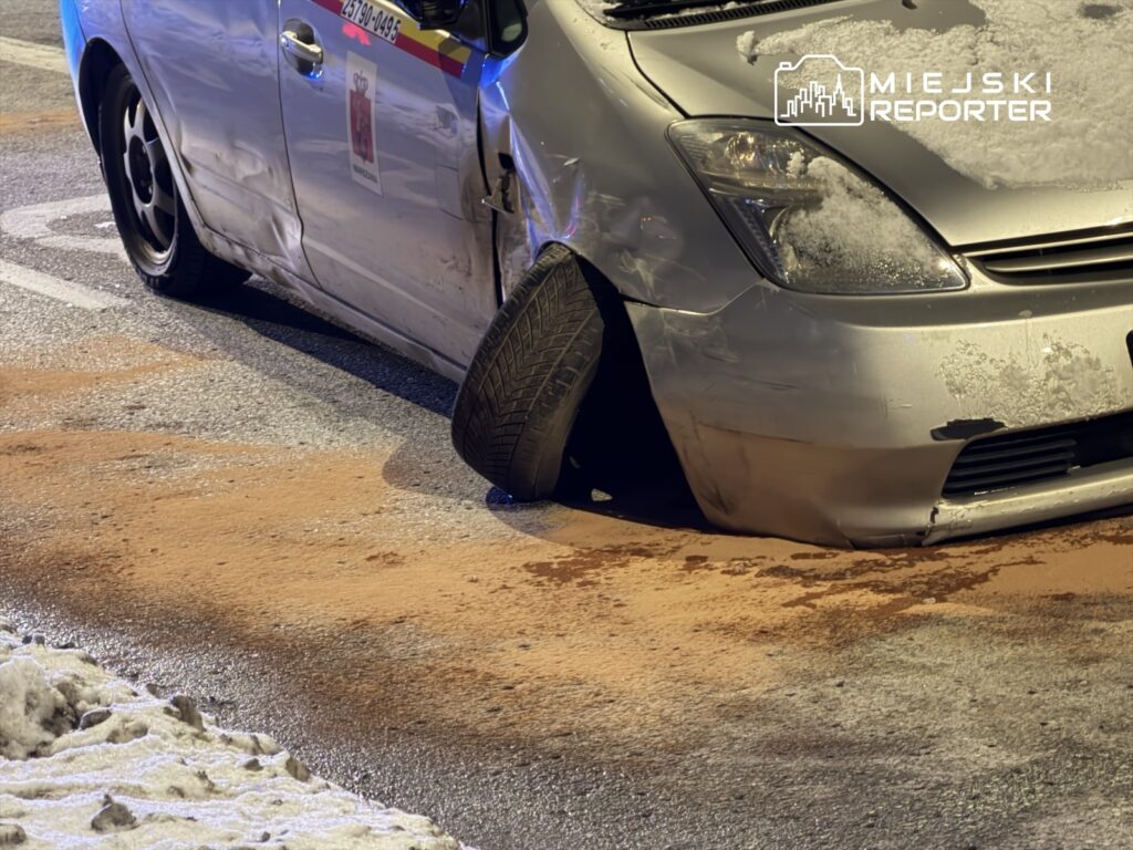 Uszkodzony samochód osobowy z opadniętym kołem stoi na zaśnieżonej ulicy, wokół widoczne plamy płynu.