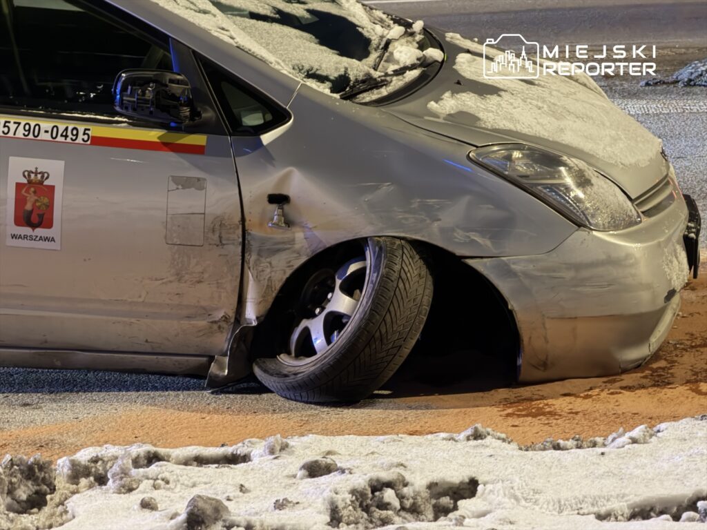 Uszkodzone taksówki z wykrzywionym kołem na śnieżnej ulicy, w tle widoczne ślady opon na drodze.