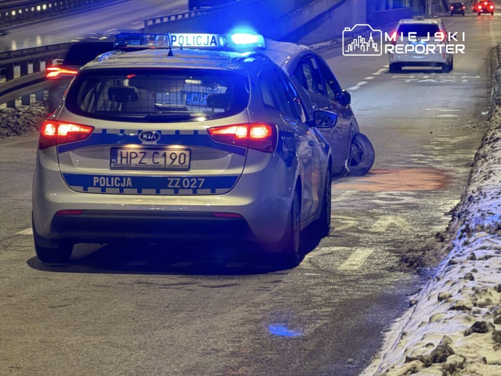 Policyjny radiowóz z włączonymi sygnałami świetlnymi zatrzymany obok uszkodzonego samochodu na ośnieżonej drodze.