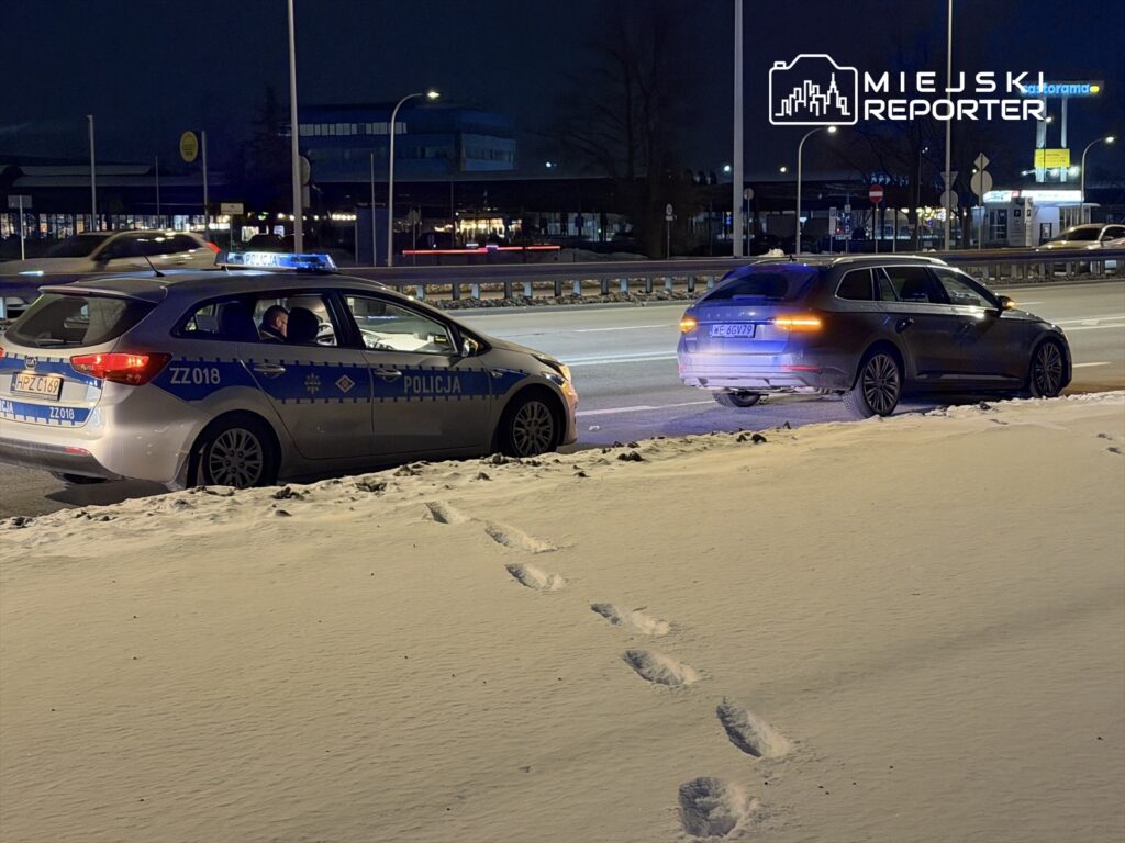 Policyjny radiowóz zatrzymany na poboczu drogi, obok stoi samochód osobowy, w tle widoczna droga i oświetlone budynki.