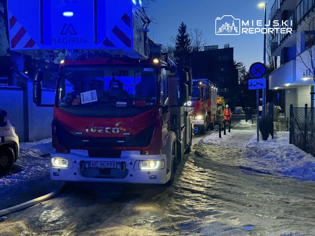 Czerwony wóz strażacki z włączonymi sygnałami świetlnymi stoi na zaśnieżonej ulicy, obok niego strażacy w odblaskowych kamizelkach.