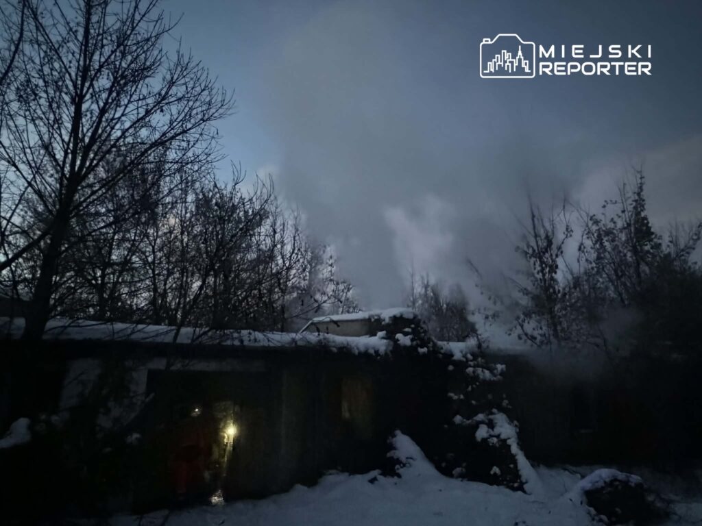 Ratownicy w odzieży ochronnej przeszukują zarywający się budynek pokryty śniegiem, w tle widoczne kłęby dymu.