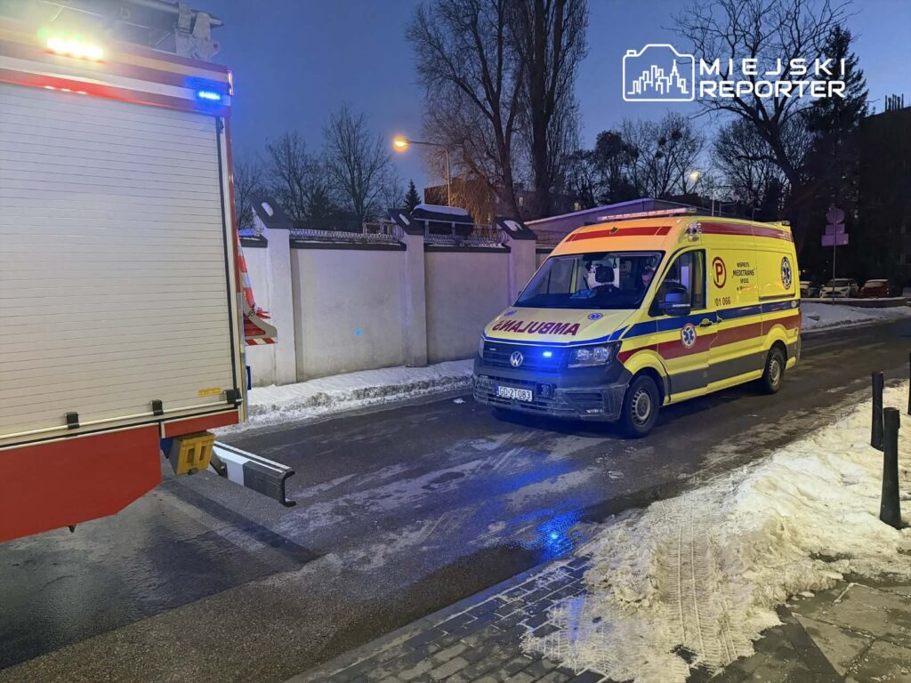 Karetka pogotowia z włączonymi sygnalizatorami stoi na ośnieżonej ulicy w mieście, obok ogrodzenia.