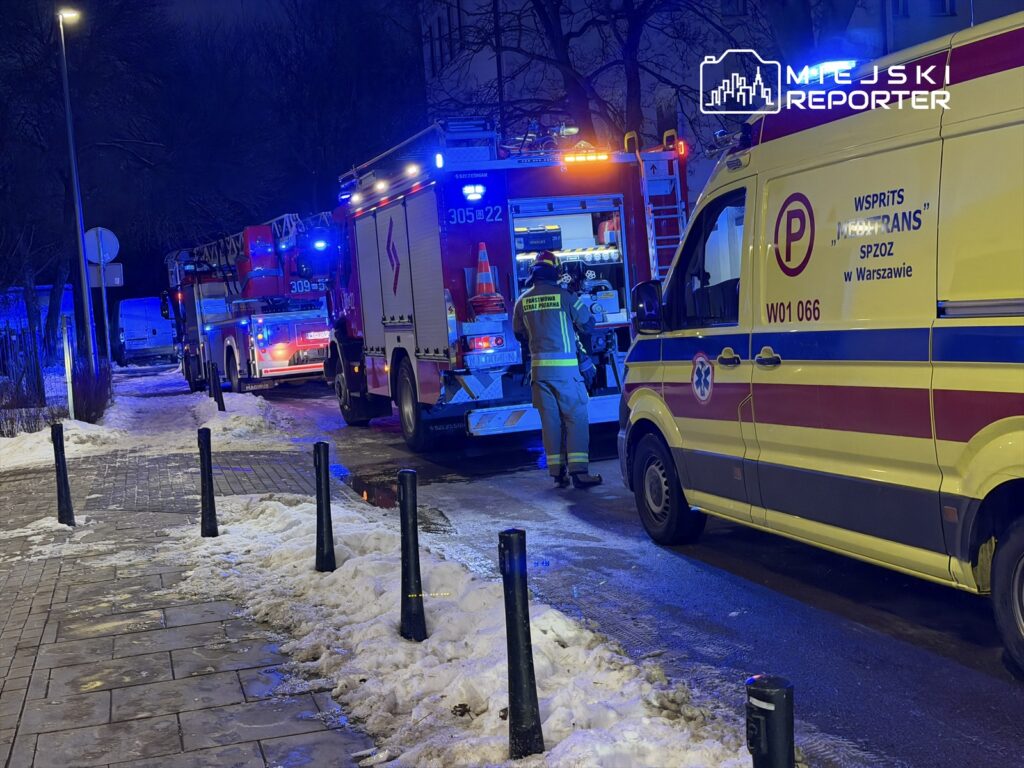 Strażacy w niebieskich mundurach pracują przy wozie strażackim na oświetlonej ulicy, obok karetki pogotowia.