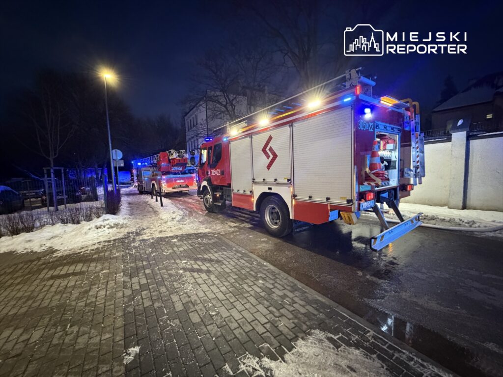Czerwony wóz strażacki z włączonymi sygnałami świetlnymi stoi na oświetlonej ulicy w zimowej scenerii.