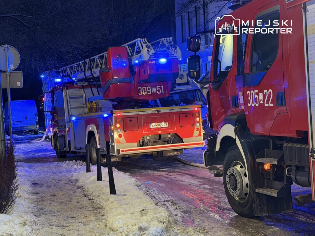 W nocy, na ośnieżonej ulicy, dwa czerwone wozy strażackie z włączonymi sygnałami świetlnymi przygotowują się do akcji.