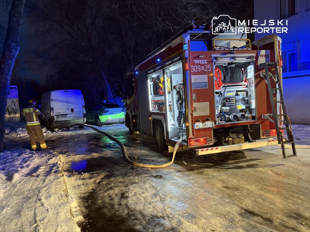 Strażak w mundurze obsługuje wąż strażacki przy zaparkowanych samochodach w oświetlonym nocą otoczeniu.