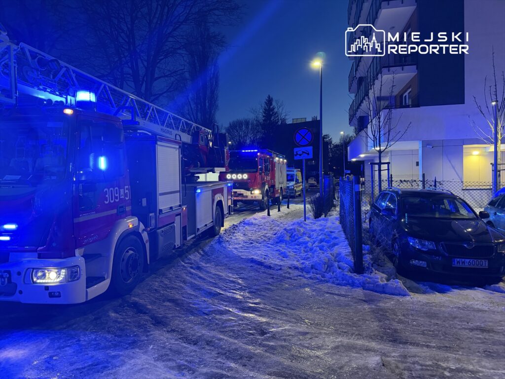 Straż pożarna z włączonymi sygnałami świetlnymi parkuje na zaśnieżonej ulicy w pobliżu nowoczesnego budynku mieszkalnego.