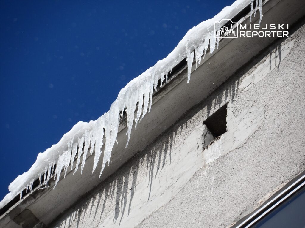 Sople lodu zwisające z krawędzi dachu budynku na tle błękitnego nieba.