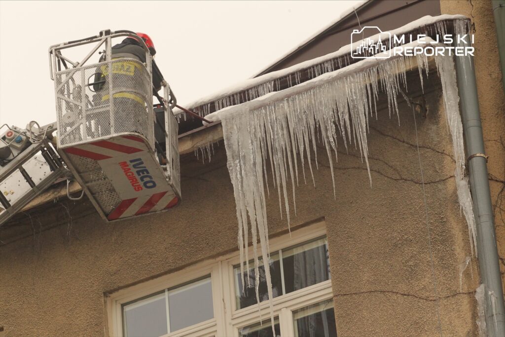 Strażak w koszu podnośnika usuwa sople lodu z krawędzi dachu budynku w pochmurny dzień.
