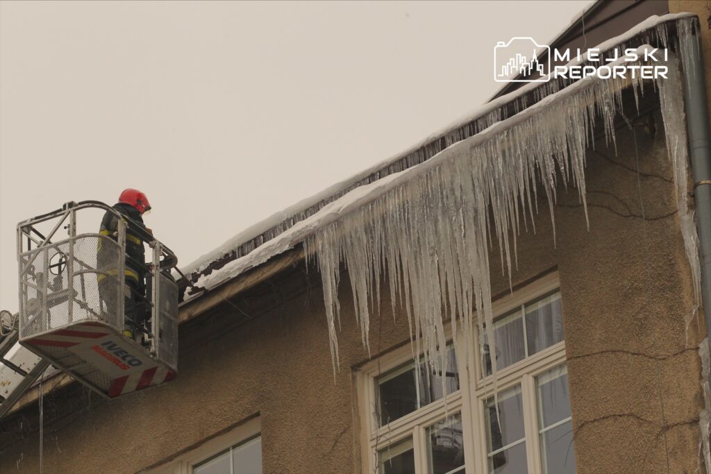 Strażak w kasku, stojący na podnośniku, usuwa sople lodu z dachu budynku w zimowej scenerii.