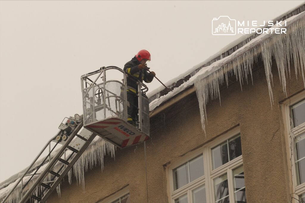 Strażak w kasku, na podnośniku, usuwa sople lodu z dachu budynku w zimowej scenerii.